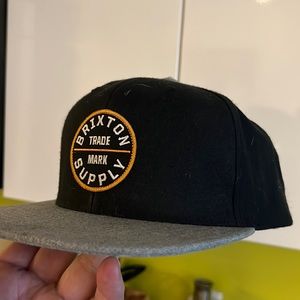 Brixton hat black with gray bill. Logo says Brixton trade mark supply. Snap back